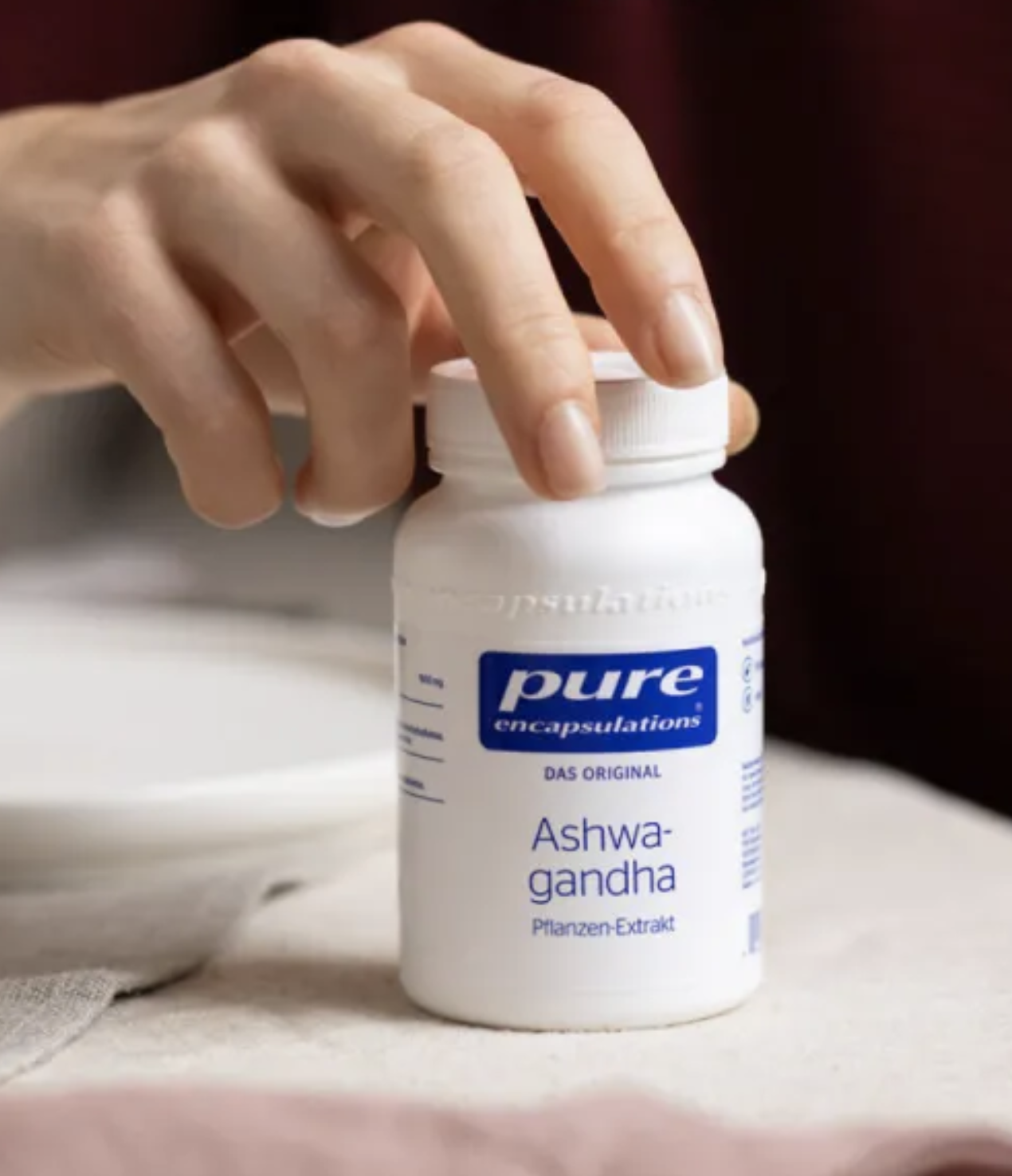 A hand reaches for a white bottle labeled "pure encapsulations Ashwagandha Pflanzen-Extrakt" on a tabletop.