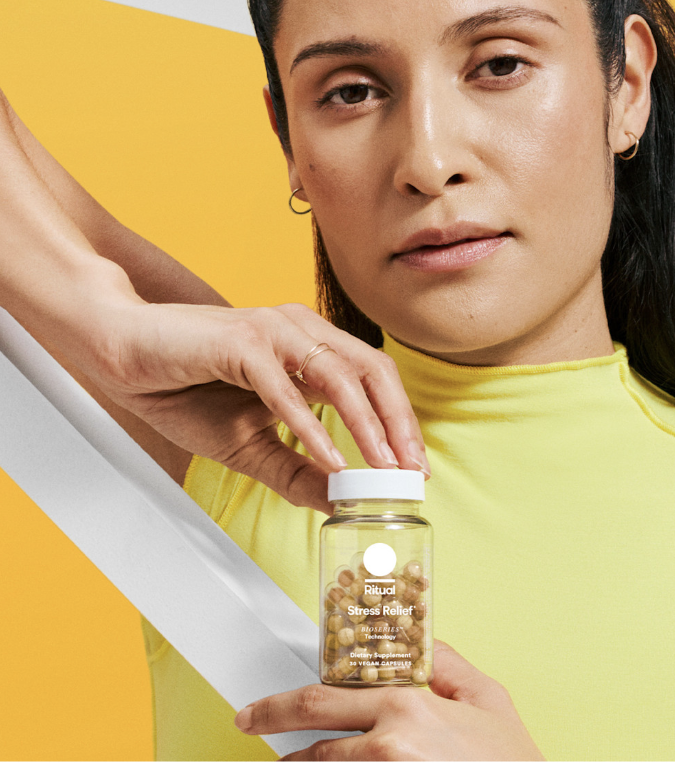 A person in a yellow top holds a clear bottle labeled "Ritual Stress Relief" capsules against a yellow and white background.