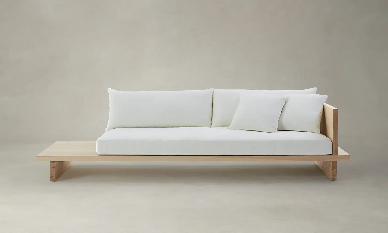 A minimalist wooden sofa with light-colored cushions and an extended flat side platform, set against a plain neutral background.