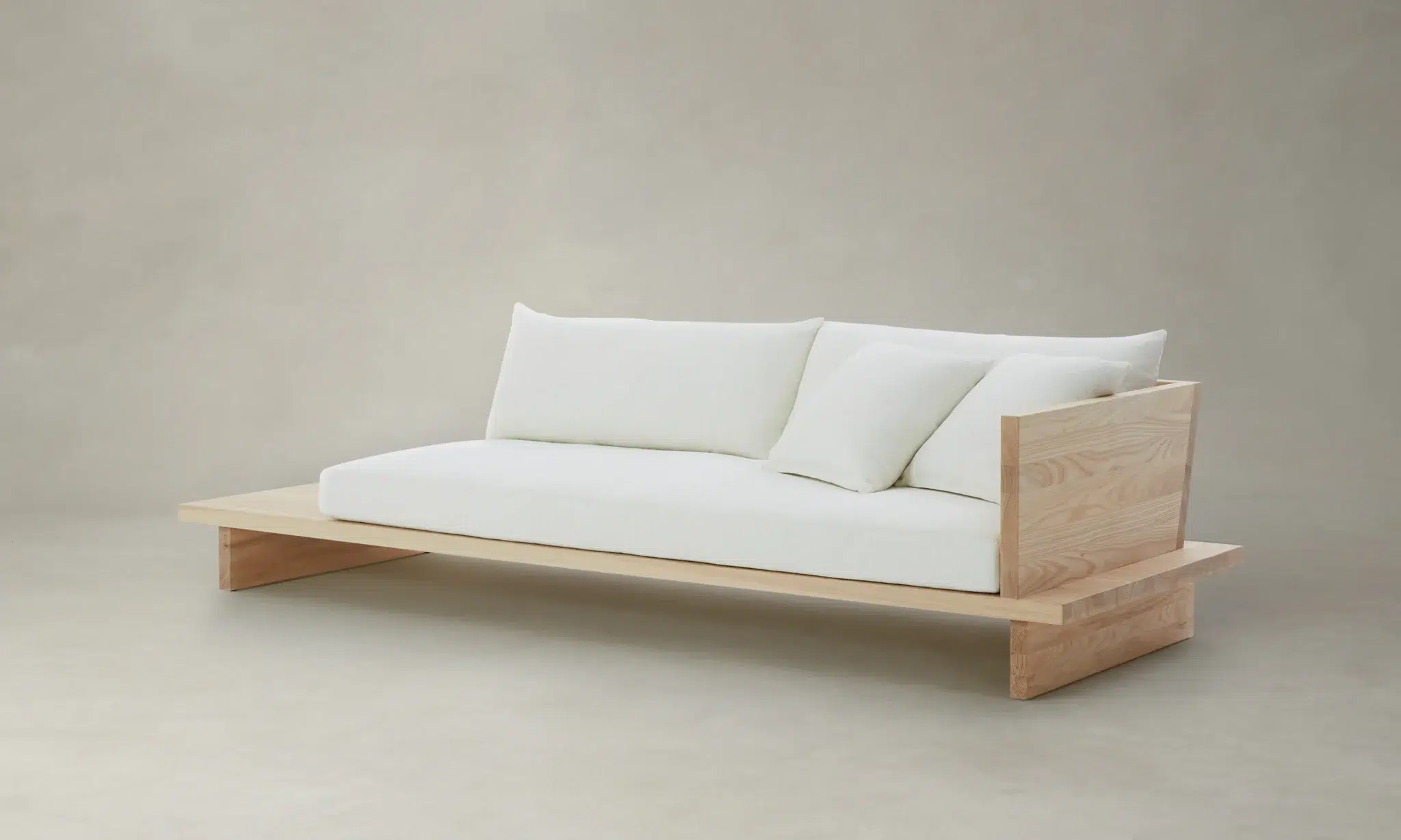 A minimalist wooden sofa with a low-profile frame, white seat cushions, and three white pillows, set against a plain light-colored background.