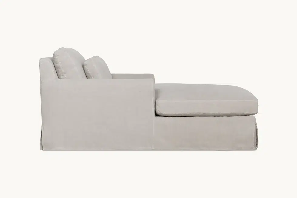 Side view of a light beige upholstered chaise lounge with two back cushions and a skirted base, set against a plain white background.
