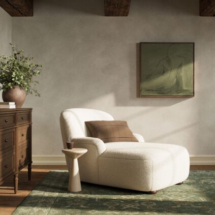 A white chaise lounge with a brown pillow sits next to a small side table in a cozy room with a dresser, potted plant, green rug, and abstract wall art.