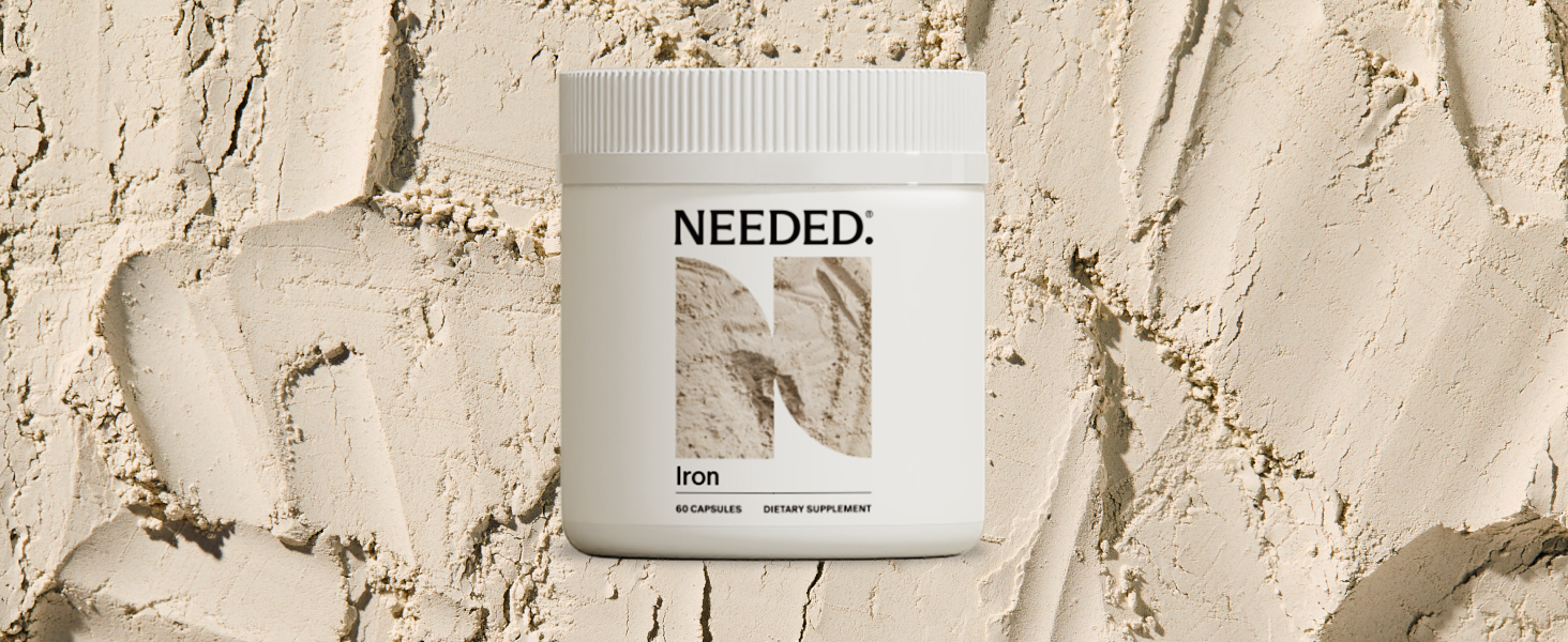 A white container labeled "Needed Iron" stands against a textured beige background that appears similar to powdered or sandy material.