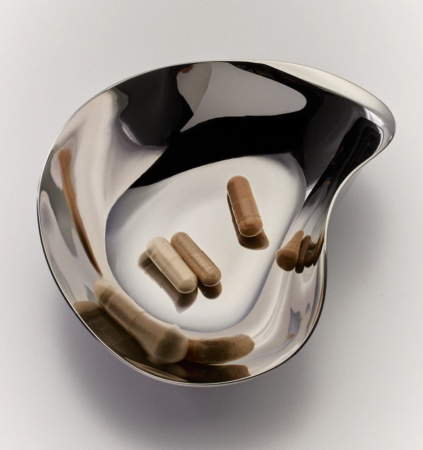 A reflective, kidney-shaped metal dish contains three beige capsules and two brown capsules, all scattered apart on the dish’s surface.