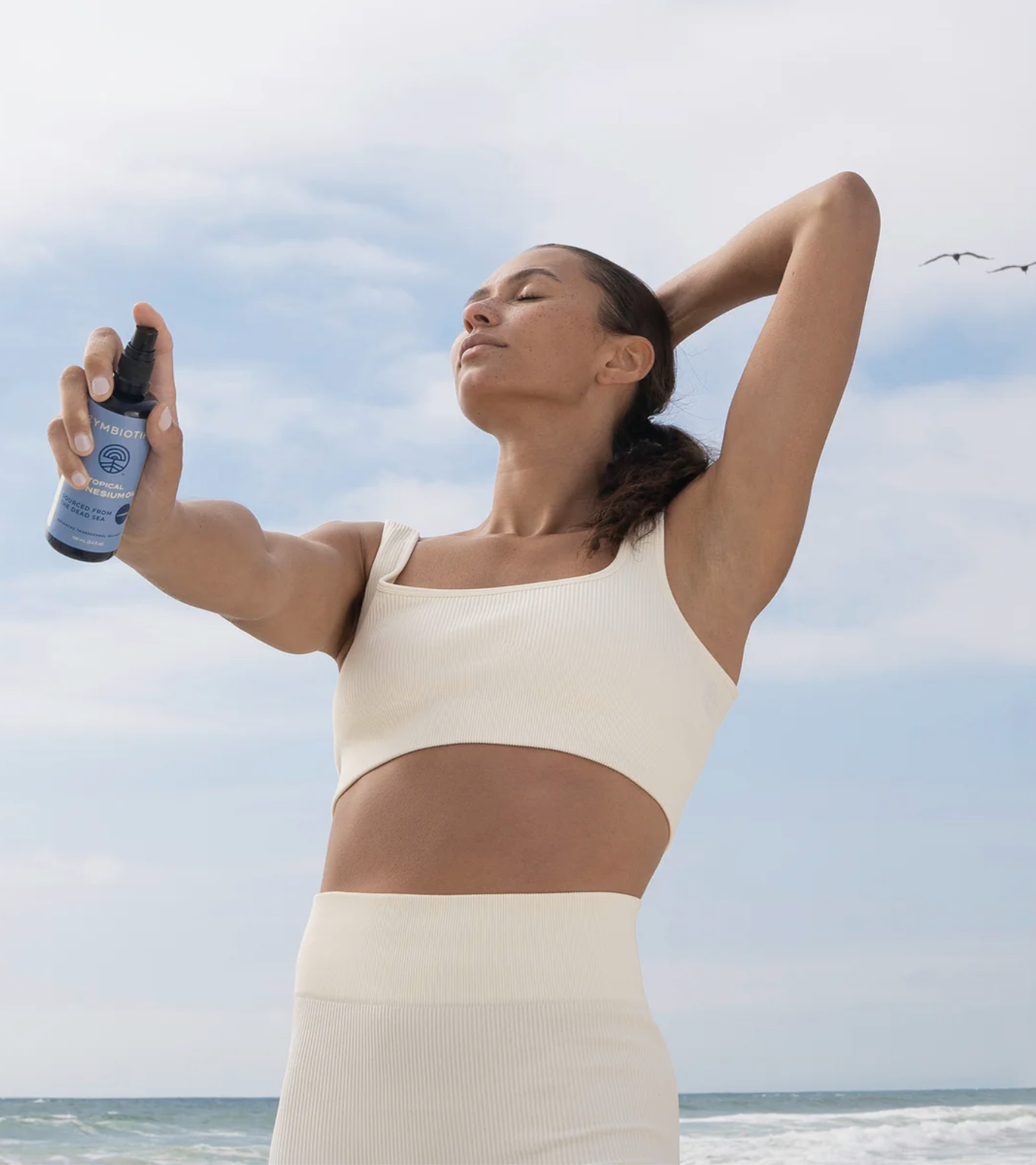 A woman in a cream sports outfit sprays a bottle on her face while standing outdoors on a beach with a blue sky in the background.