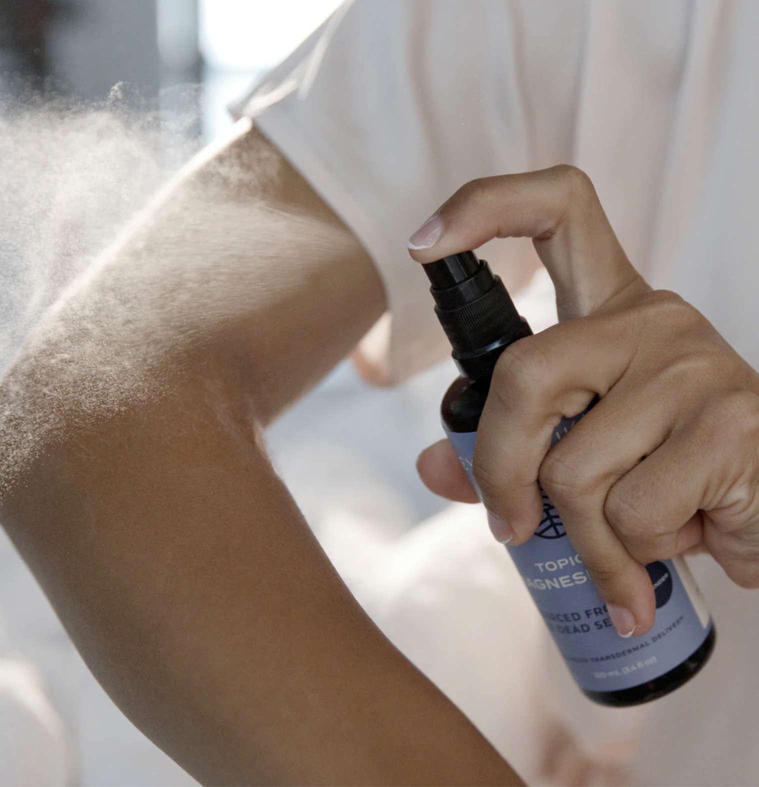 A person spraying a mist from a bottle onto their forearm, with the label on the bottle partially visible.