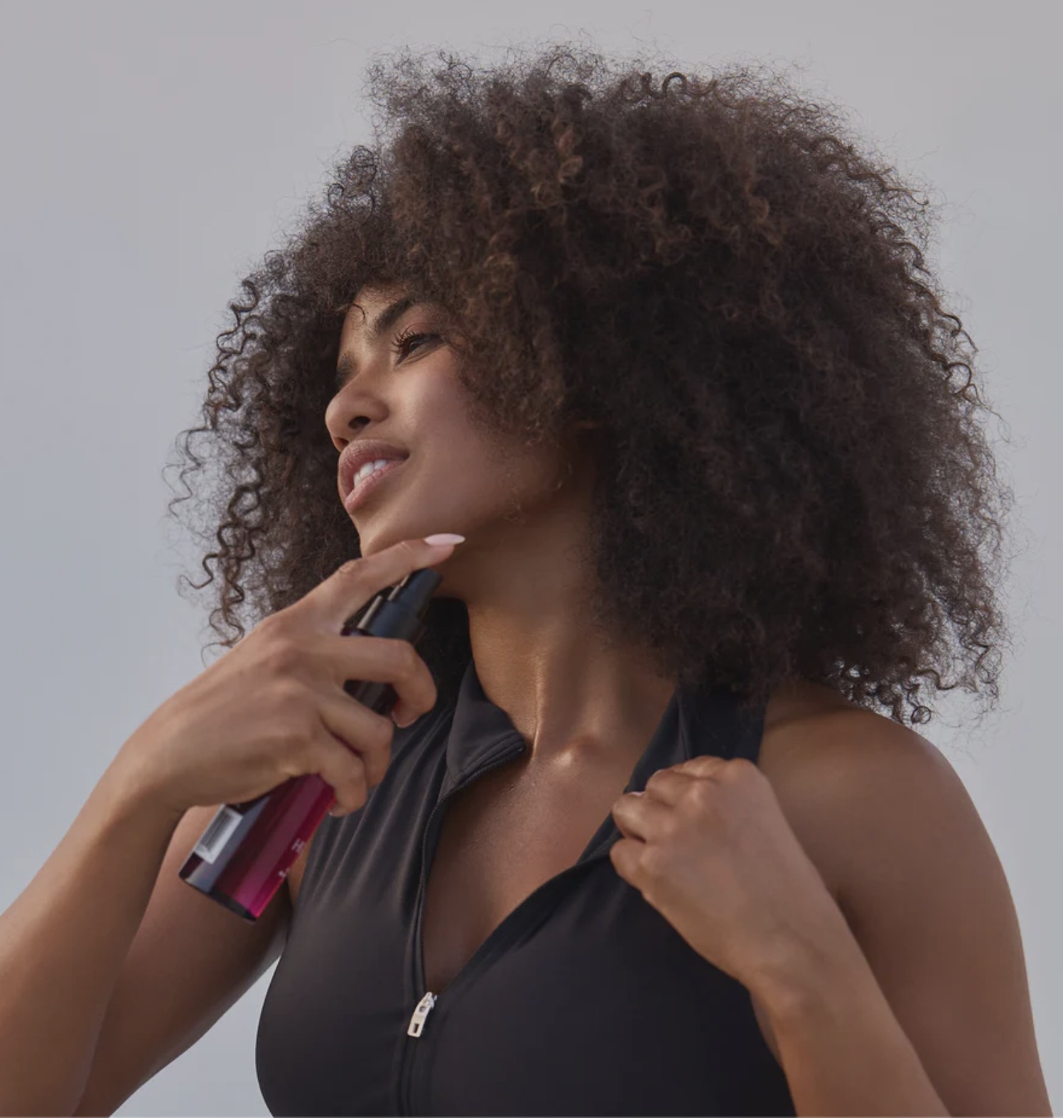 A woman with curly hair sprays a bottle onto her neck while wearing a sleeveless black zip-up top.