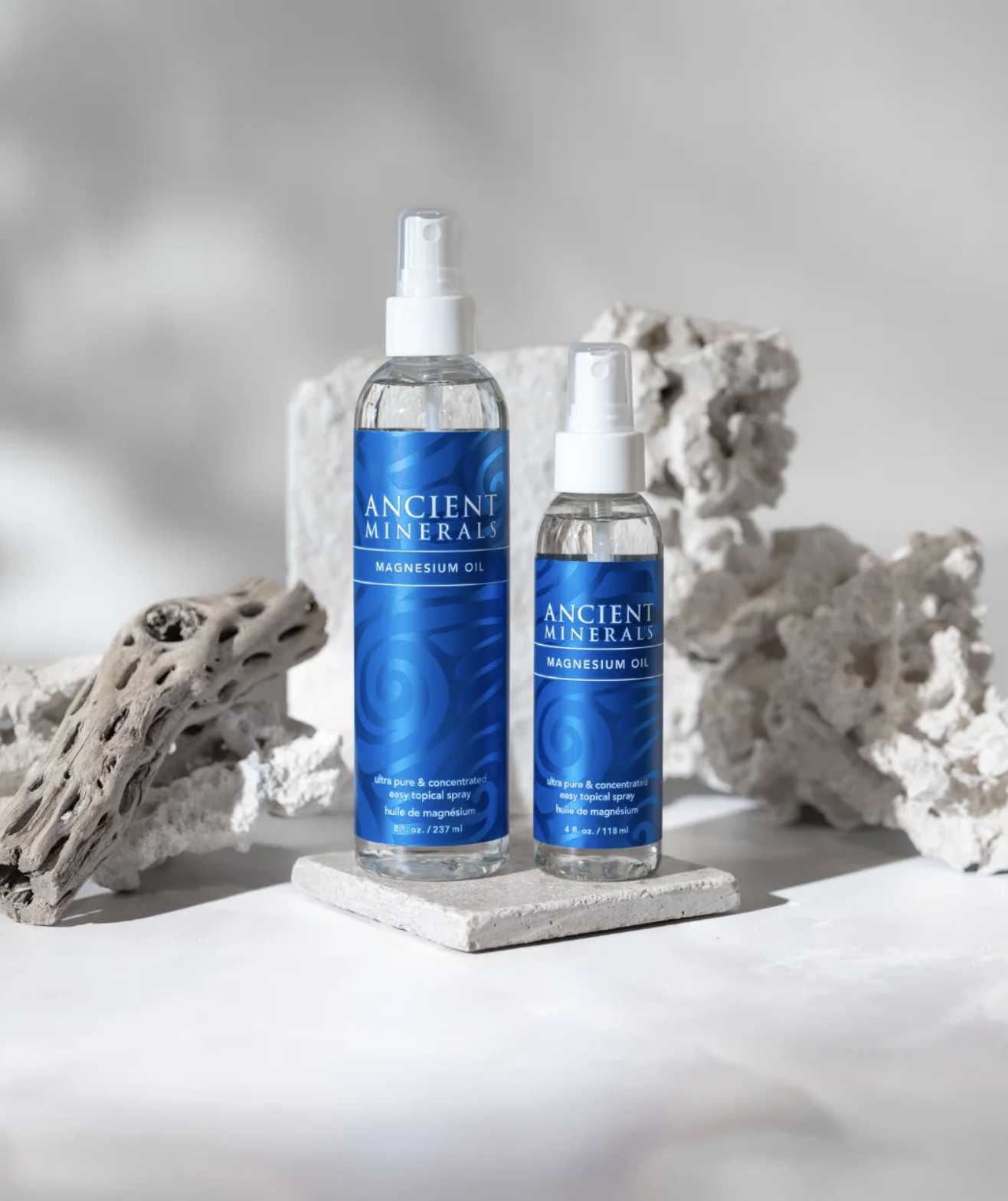 Two bottles of Ancient Minerals Magnesium Oil spray are displayed on white stone slabs with textured rocks in the background.
