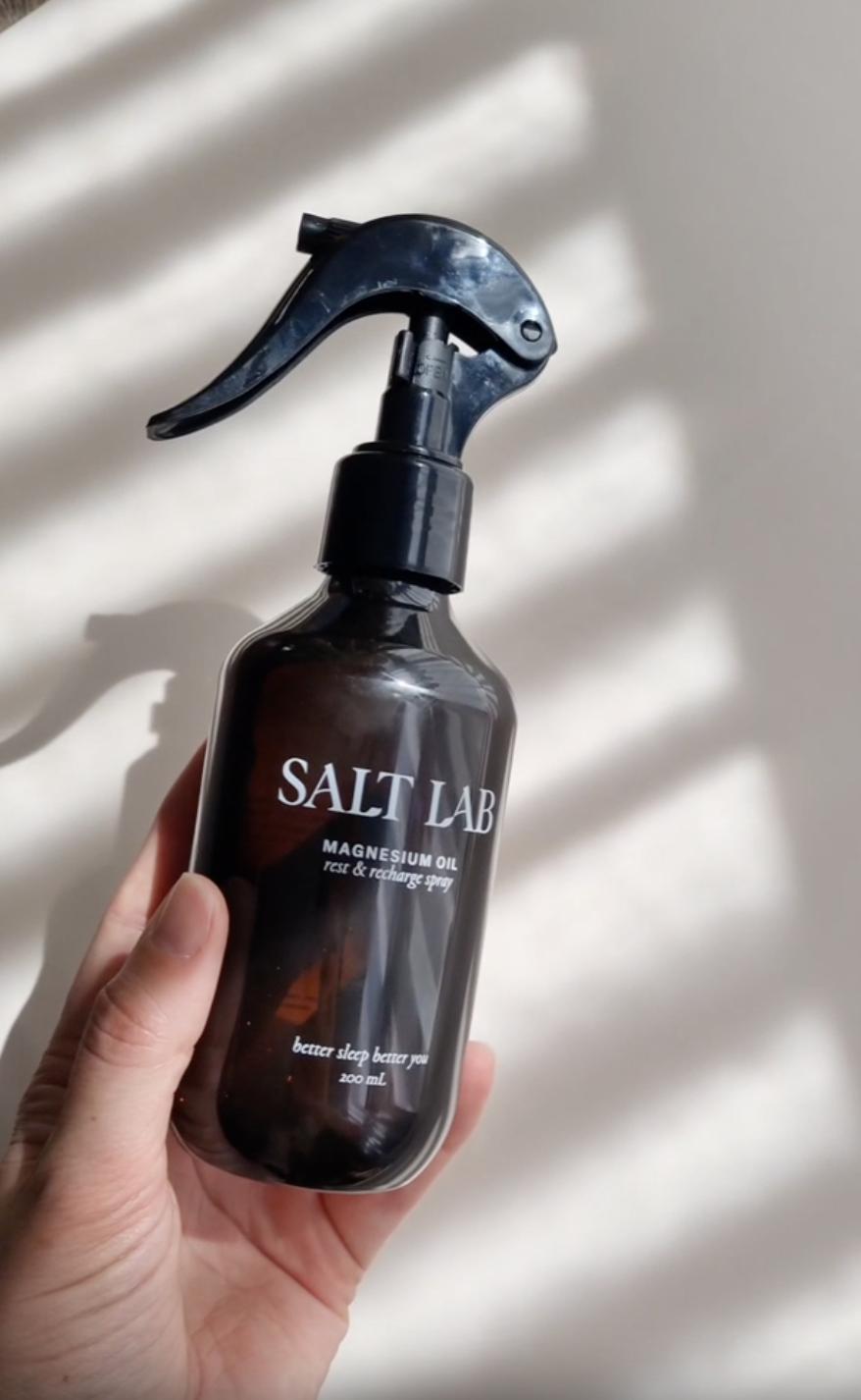 A hand holds a black spray bottle labeled "Salt Lab Magnesium Oil" against a white background with striped shadows.