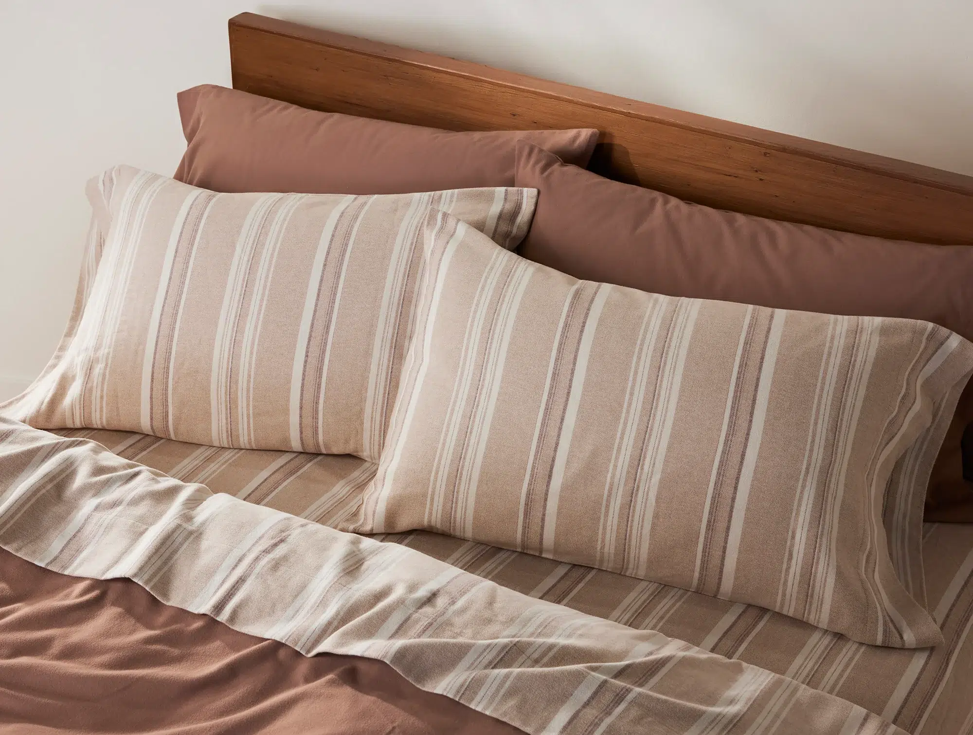 A made bed with a wooden headboard, two striped pillows, a striped sheet, and solid brown pillowcases and fitted sheet.