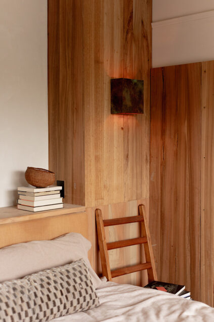 A cozy bedroom corner with wood-paneled walls, a bed with a patterned pillow, a wooden chair, stacked books, and a small basket. Warm light glows from a wall fixture.