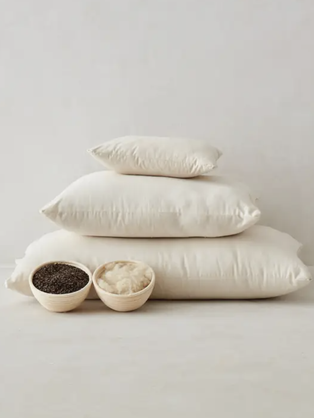 Three cream-colored pillows stacked in descending size order, with a smaller pillow on top; two small bowls in front contain black seeds and white stuffing material.