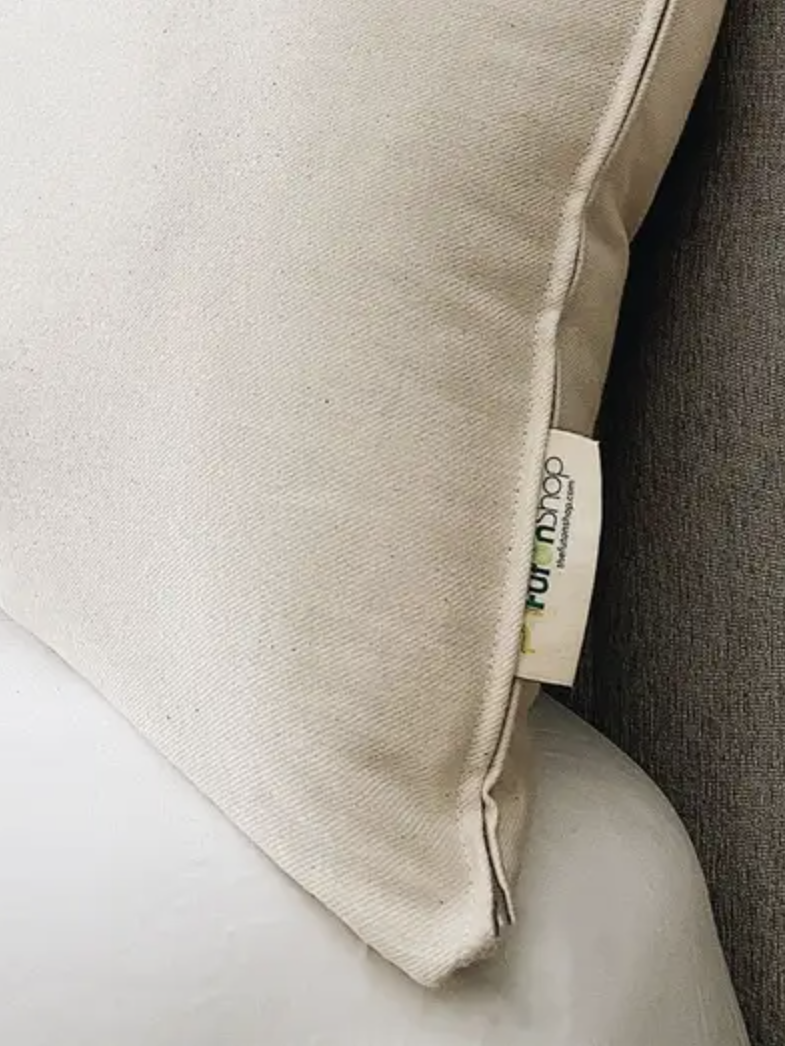 A close-up of a beige pillow with a fabric tag showing partial text, resting on a white surface next to a gray upholstered object.