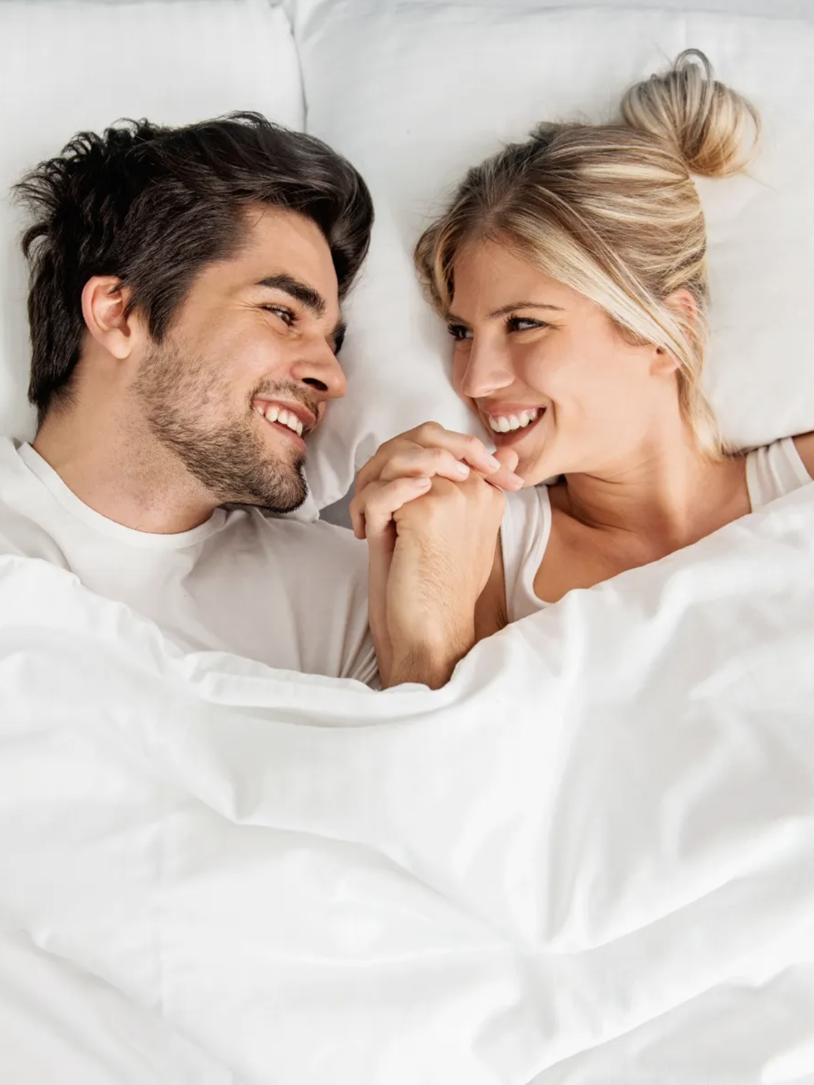 A man and woman lie in bed under white sheets, facing each other and smiling while holding hands.