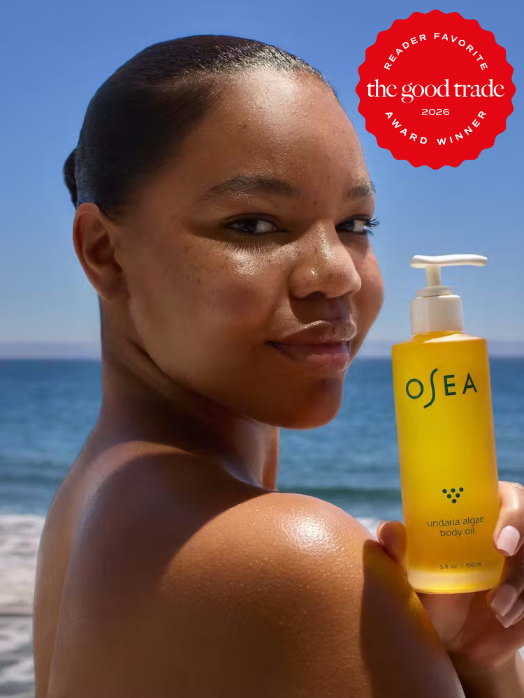 A woman stands on a beach holding a bottle of Osea Undaria Algae Body Oil. A “Reader Favorite The Good Trade Award Winner 2026” badge appears in the corner.
