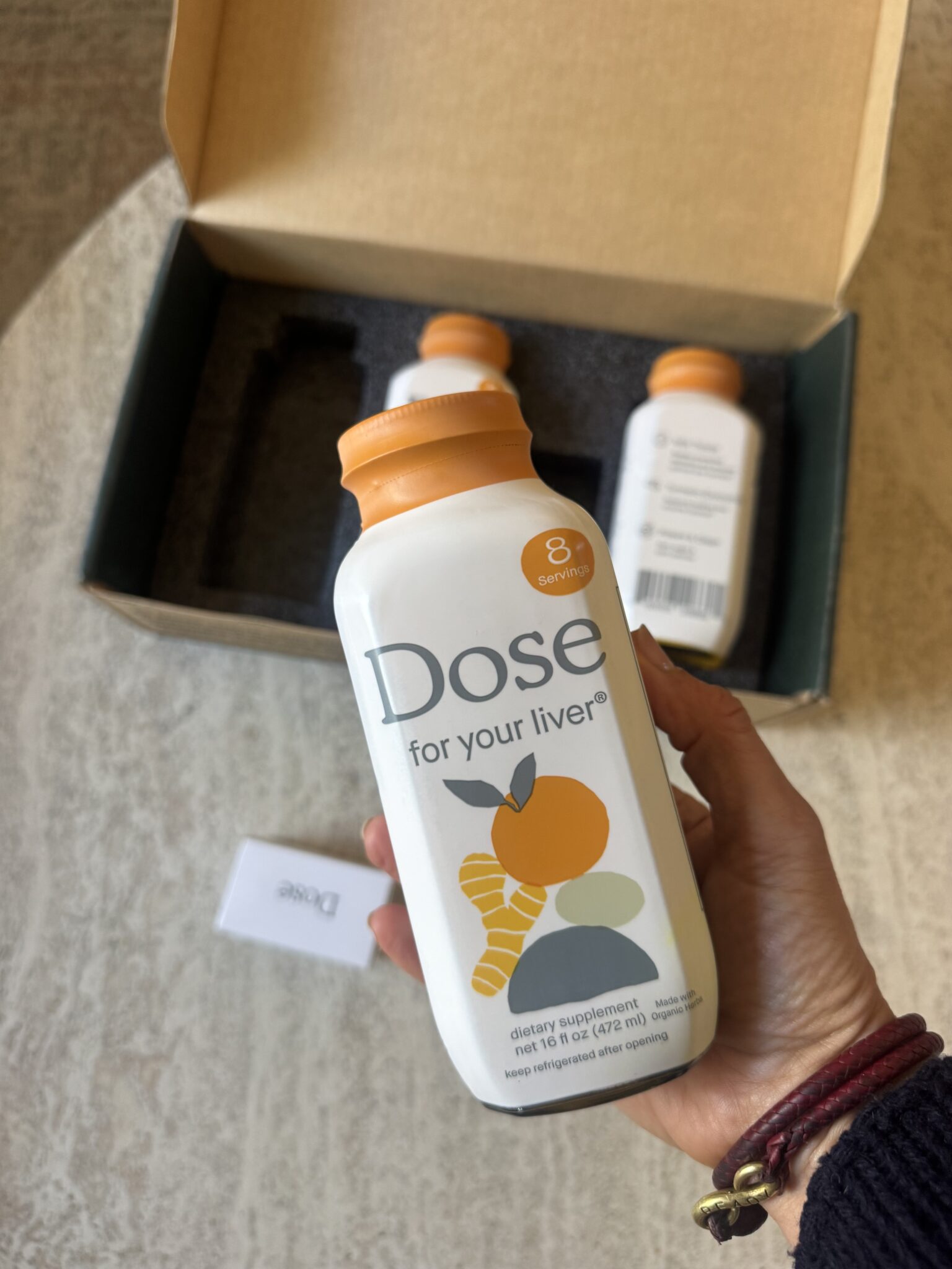 A hand holds a bottle labeled "Dose for your liver" with an orange cap; two more bottles and a box are in the background.