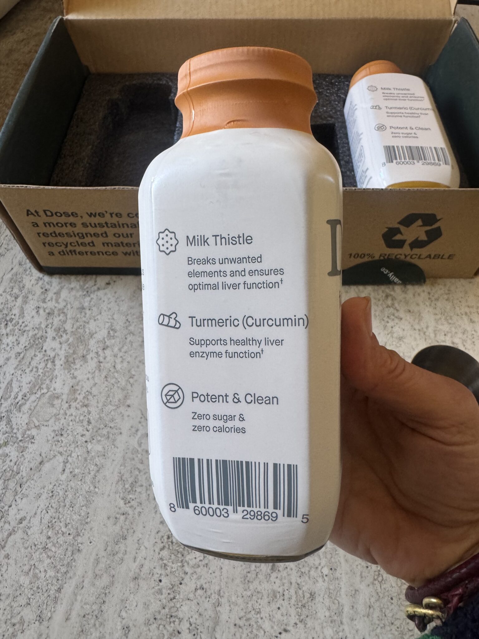 A hand holds a white bottle showing supplement ingredients: milk thistle, turmeric (curcumin), and a note about zero sugar and calories. Another bottle and cardboard box are in the background.