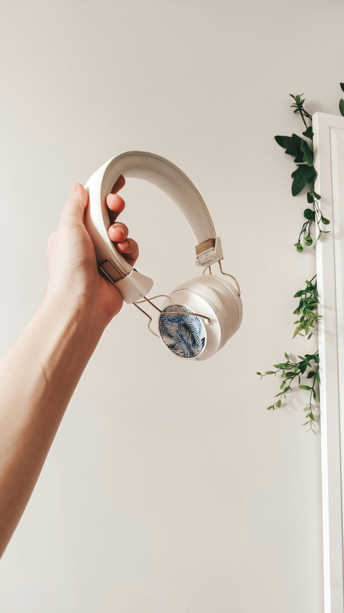 A hand holds up a pair of white over-ear headphones with a blue leaf pattern on the ear cup; green vines hang in the background.