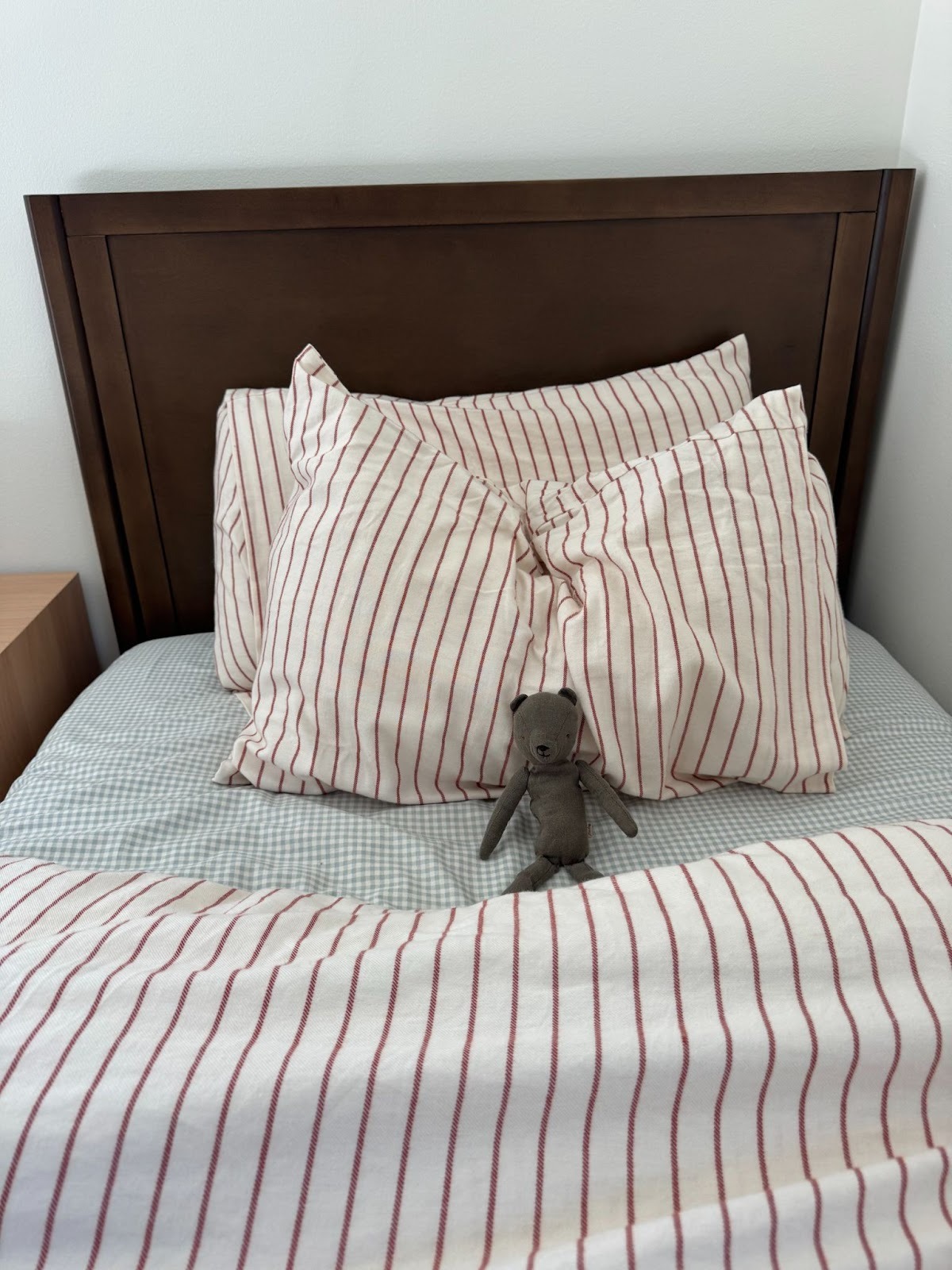 A small stuffed bear sits on a neatly made bed with a wooden headboard and red-and-white striped bedding and pillows.