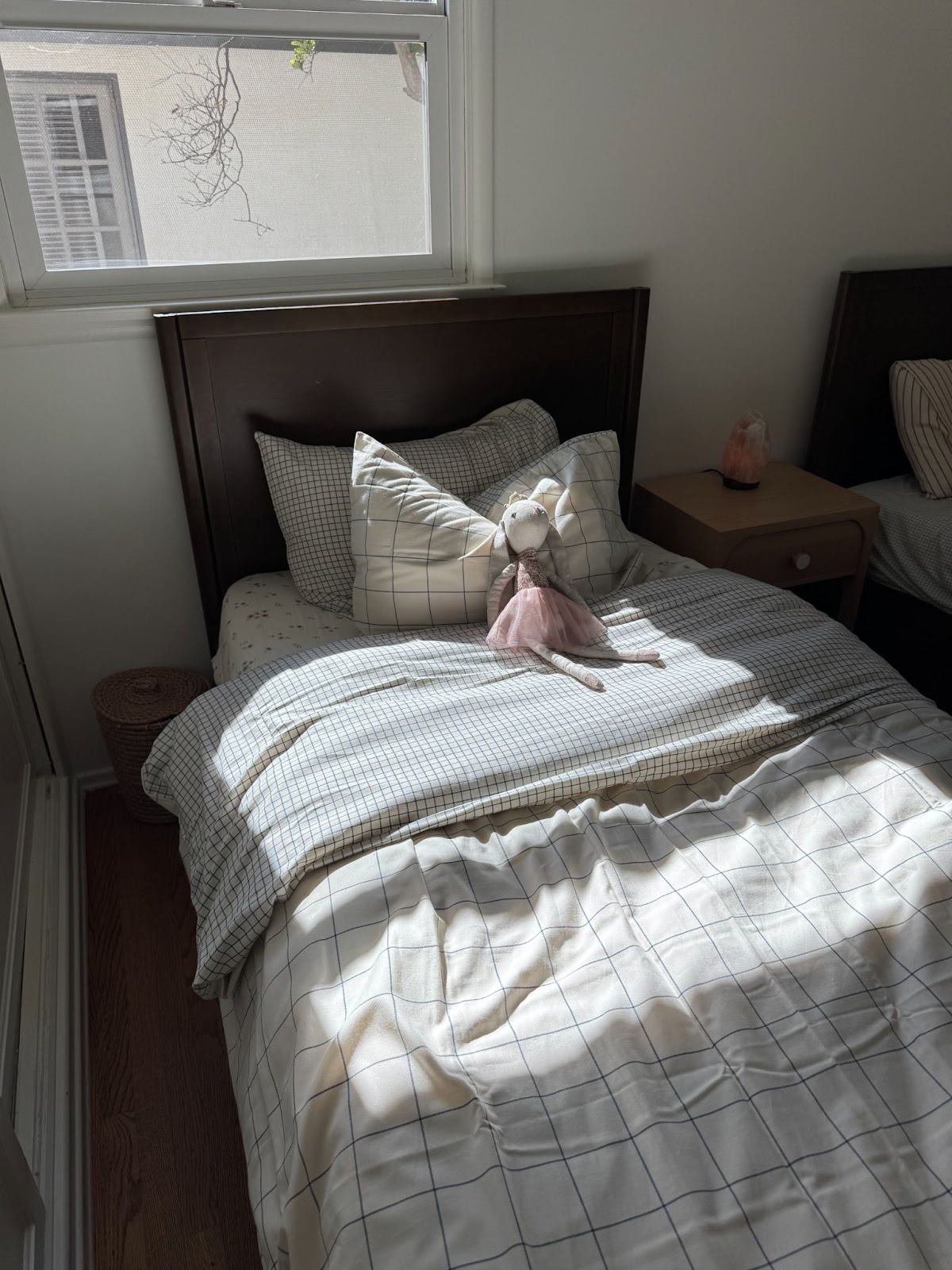 A neatly made bed with checkered bedding, a stuffed doll in a pink dress on the pillow, and sunlight streaming through a window above. A nightstand and part of another bed are visible.