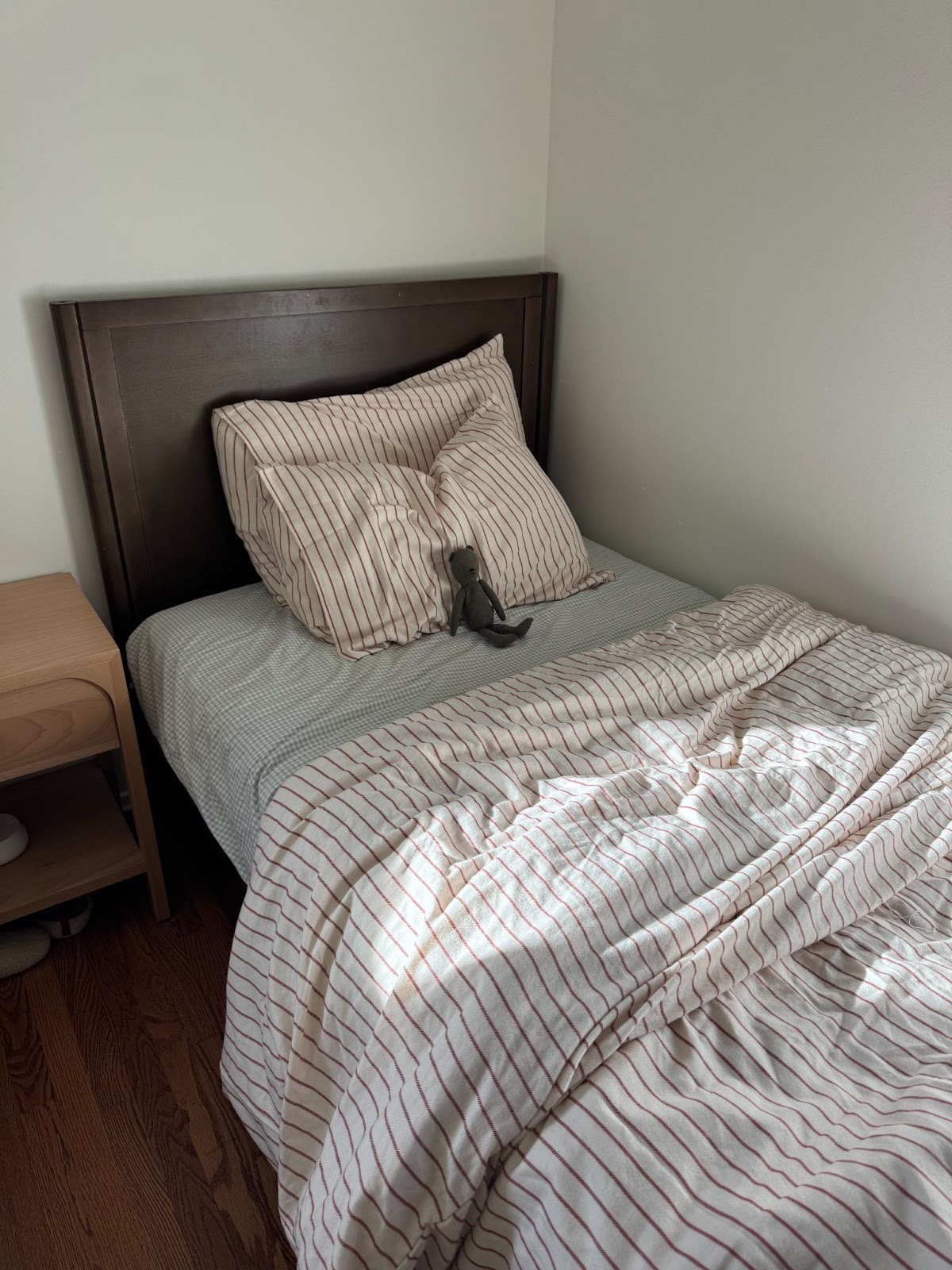 A neatly made twin bed with striped bedding and pillows, with a small stuffed animal placed in the center, next to a wooden nightstand.