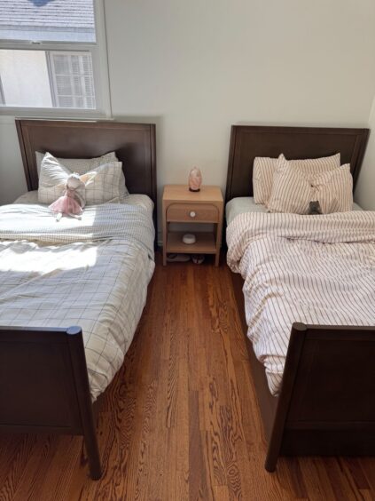 Two twin beds with wooden frames are placed side by side, separated by a small nightstand, in a sunlit bedroom with hardwood floors.