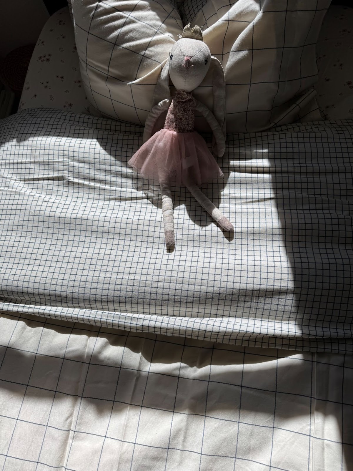 A stuffed bunny doll in a pink dress is sitting on a neatly made bed with grid-patterned sheets, illuminated by sunlight.