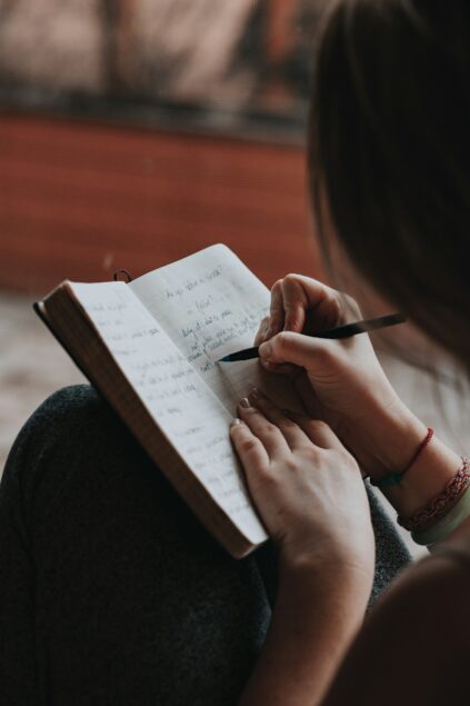 A person writes with a pen in a notebook, seated with legs crossed. Handwritten notes and a red string bracelet are visible.
