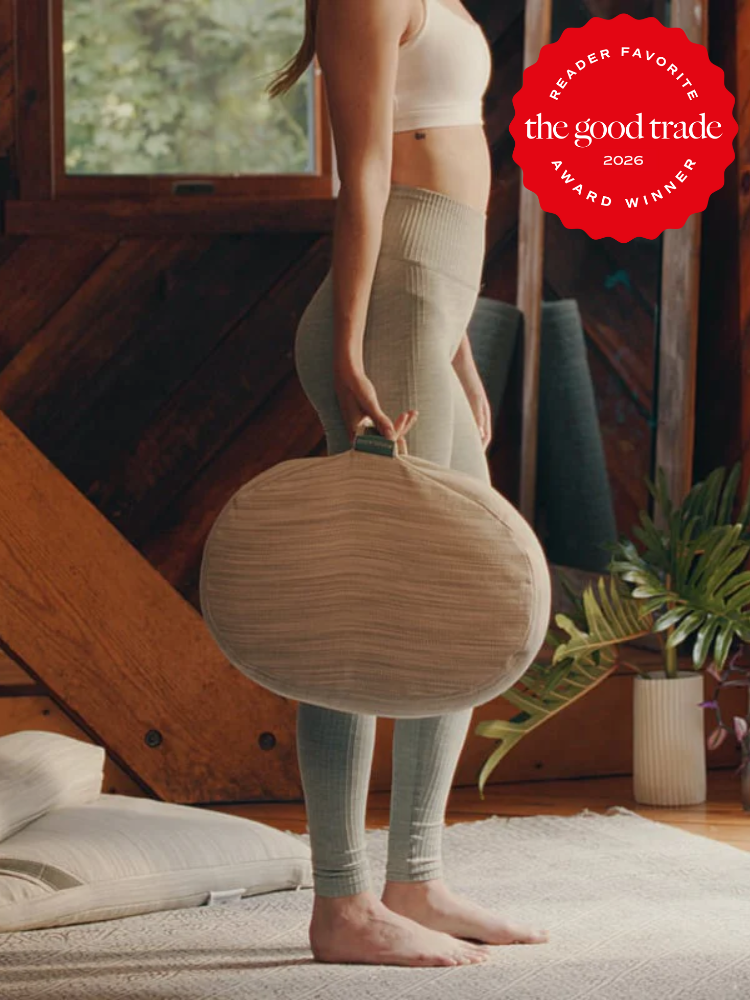 A woman in a sports bra and leggings stands indoors holding a round meditation cushion by its handle. A red award badge appears in the top right corner.