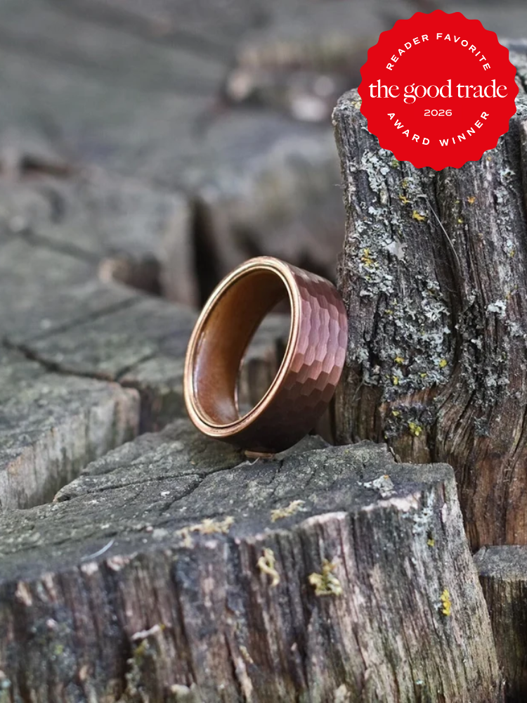 A hammered copper ring rests upright on weathered wooden tree stumps; a red "The Good Trade Reader Favorite Award Winner 2026" badge appears in the top right corner.