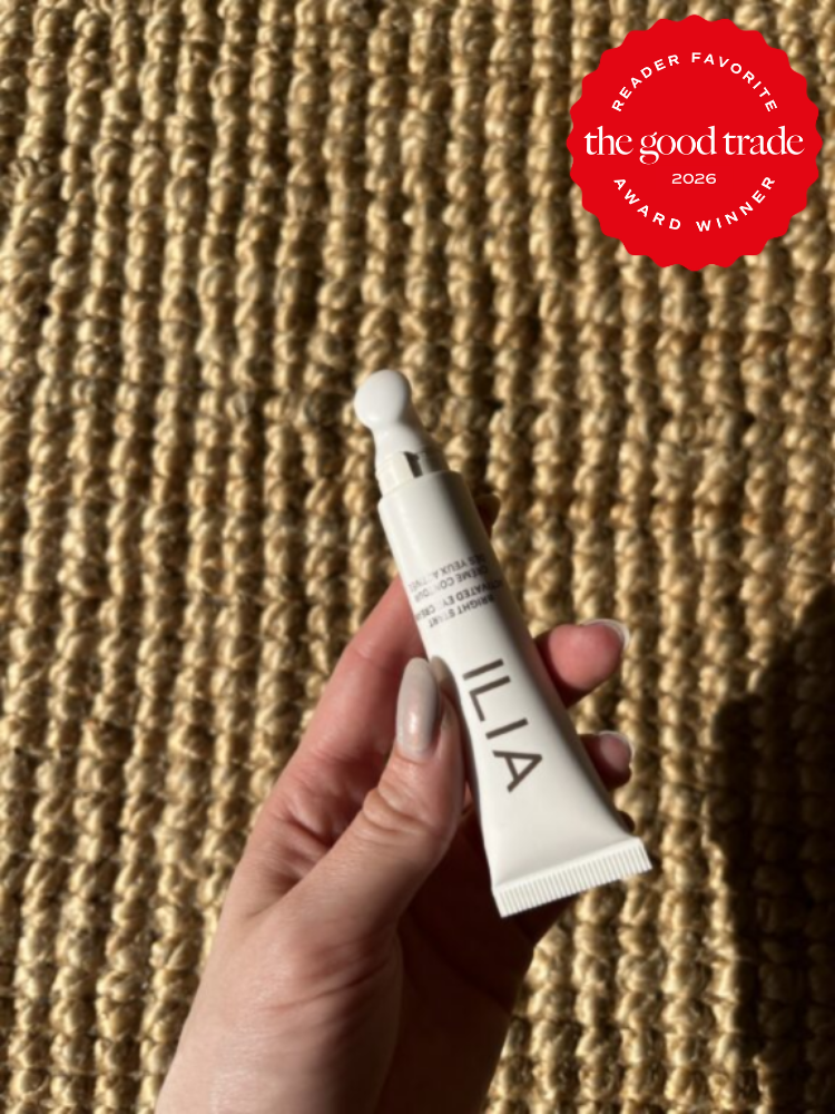A hand holds a tube of ILIA product against a woven textured background. A red badge in the corner reads “the good trade 2026 award winner.”.