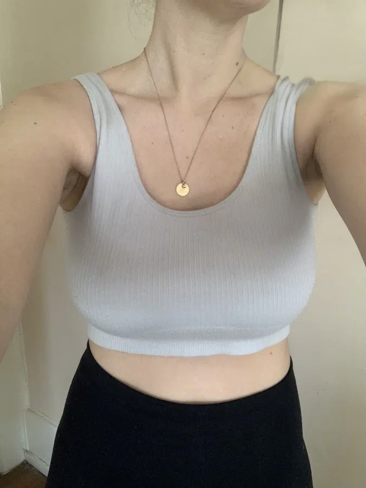 A person wearing a light gray ribbed tank top and a round pendant necklace, photographed from the neck down indoors.