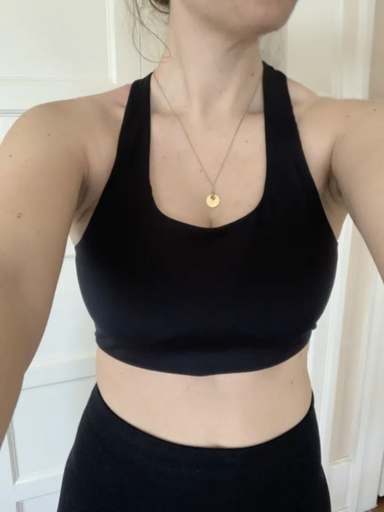 A person wearing a black sleeveless crop top and matching black bottom, with a gold pendant necklace, stands indoors.