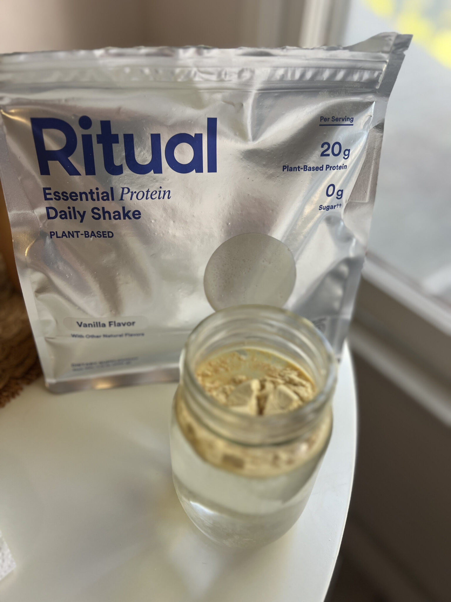 A packet of Ritual Essential Protein Daily Shake sits behind a glass jar with protein powder and water, placed on a white surface near a window.