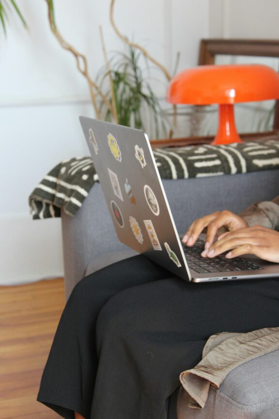 Person typing on a sticker-covered laptop while sitting on a gray chair, with an orange lamp and a plant in the background.