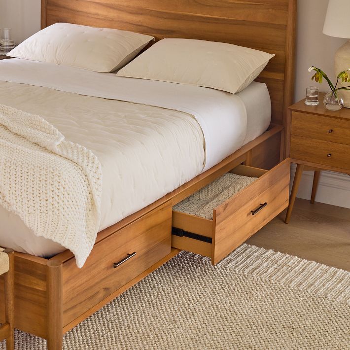 A wooden bed with white bedding features built-in storage drawers, one of which is open. A nightstand with flowers and a glass of water stands beside the bed on a textured rug.