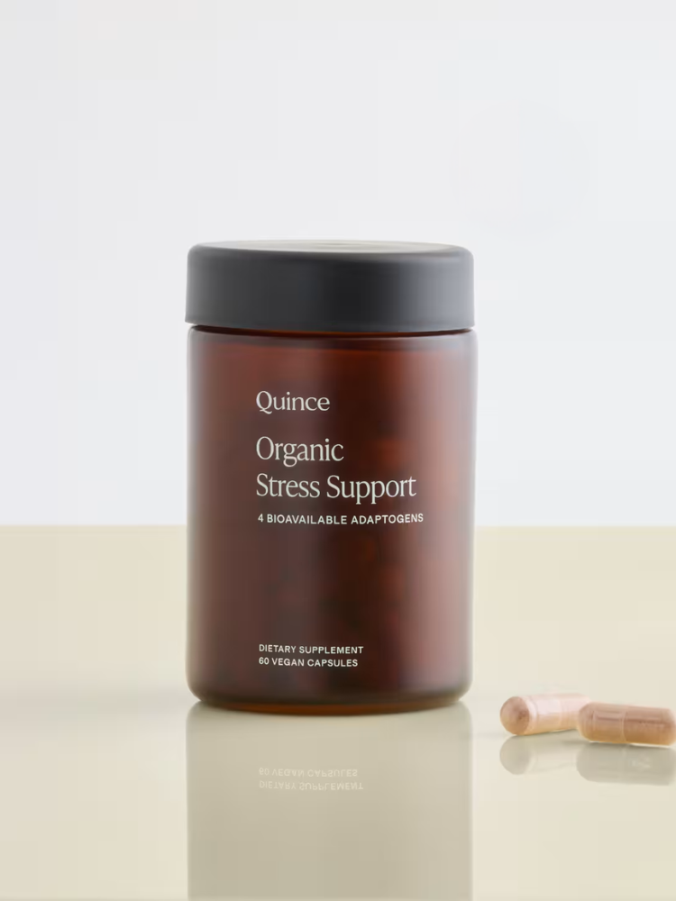 A brown jar labeled "Quince Organic Stress Support" with two capsules beside it on a neutral surface.