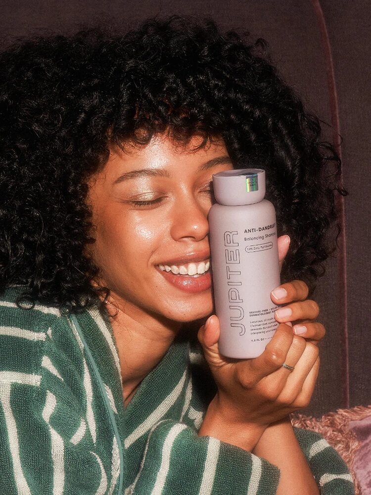 A woman in a green striped robe smiles and holds a bottle labeled "Jupiter Anti-Dandruff Shampoo" close to her face.