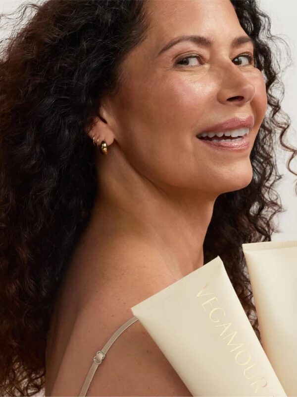 A woman with curly dark hair smiles while holding two tubes of Vegamour GRO+ Advanced hair products against a neutral background.