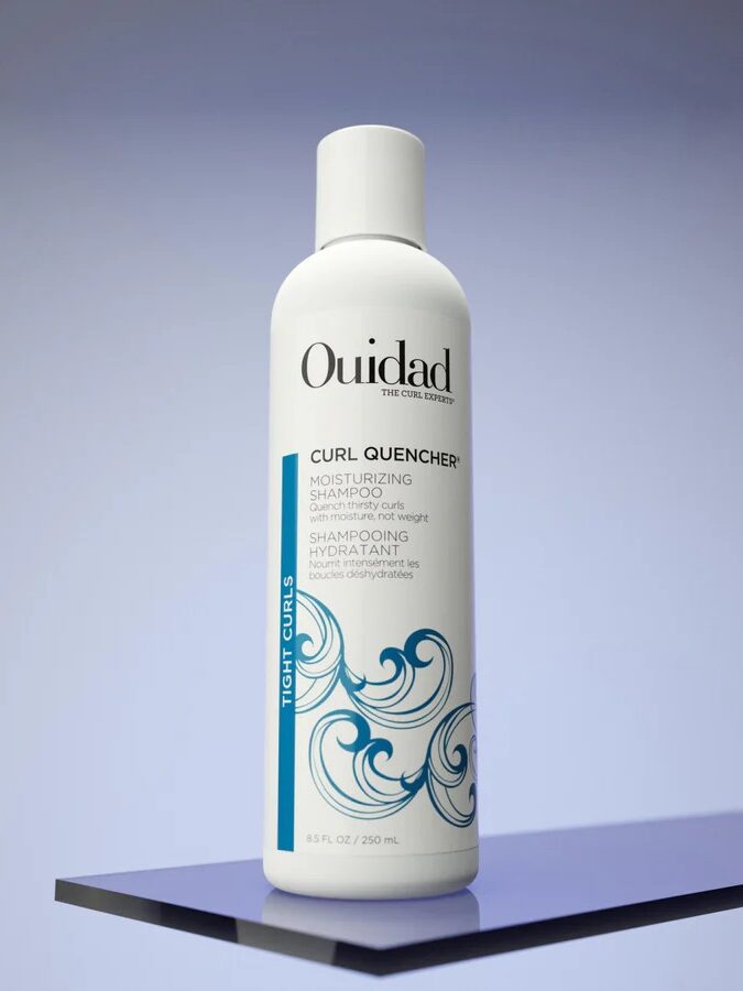 A bottle of Ouidad Curl Quencher Moisturizing Shampoo for tight curls, 8.5 fl oz, is displayed upright on a reflective surface with a light blue background.