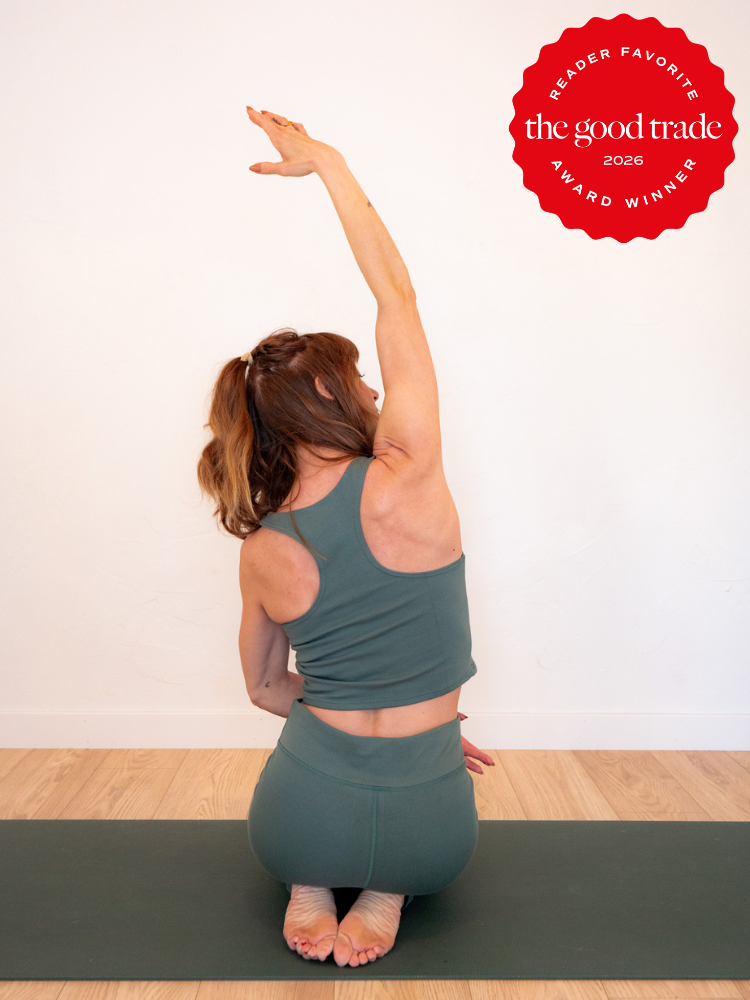 A woman in green activewear sits on a yoga mat, kneeling with one arm raised overhead in a side stretch. A red "The Good Trade 2026 Award Winner" badge is in the top right corner.