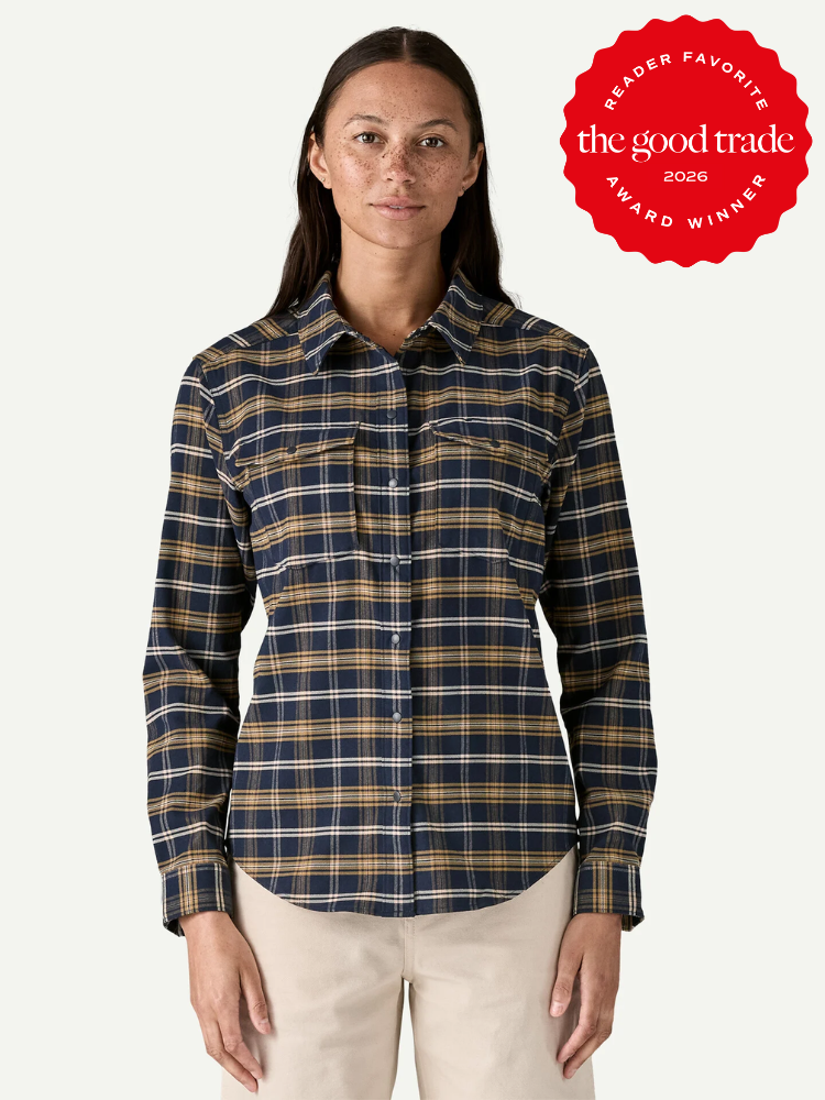 A woman wearing a navy and yellow plaid button-up shirt stands facing forward. A red "The Good Trade 2026 Award Winner" badge appears in the top right corner.