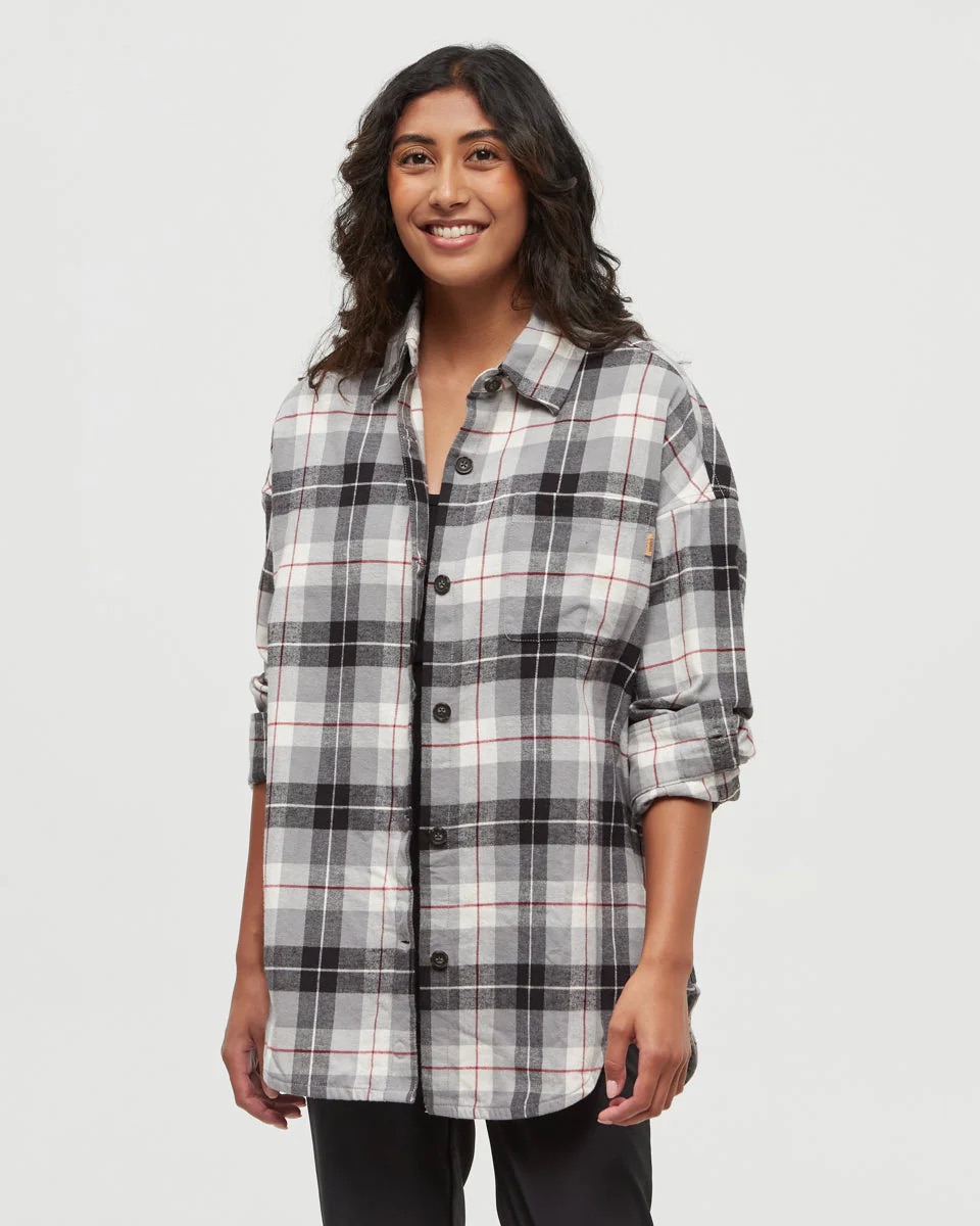 A person stands smiling, wearing a black, white, and gray plaid button-up shirt with sleeves rolled up, against a plain light background.
