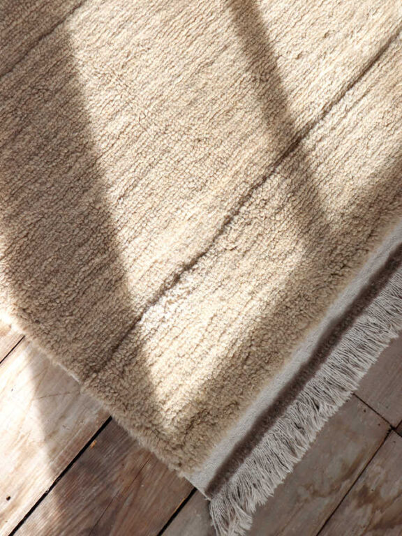 A beige textured rug with fringed edges lies on a wooden floor, with sunlight and shadows falling across its surface.