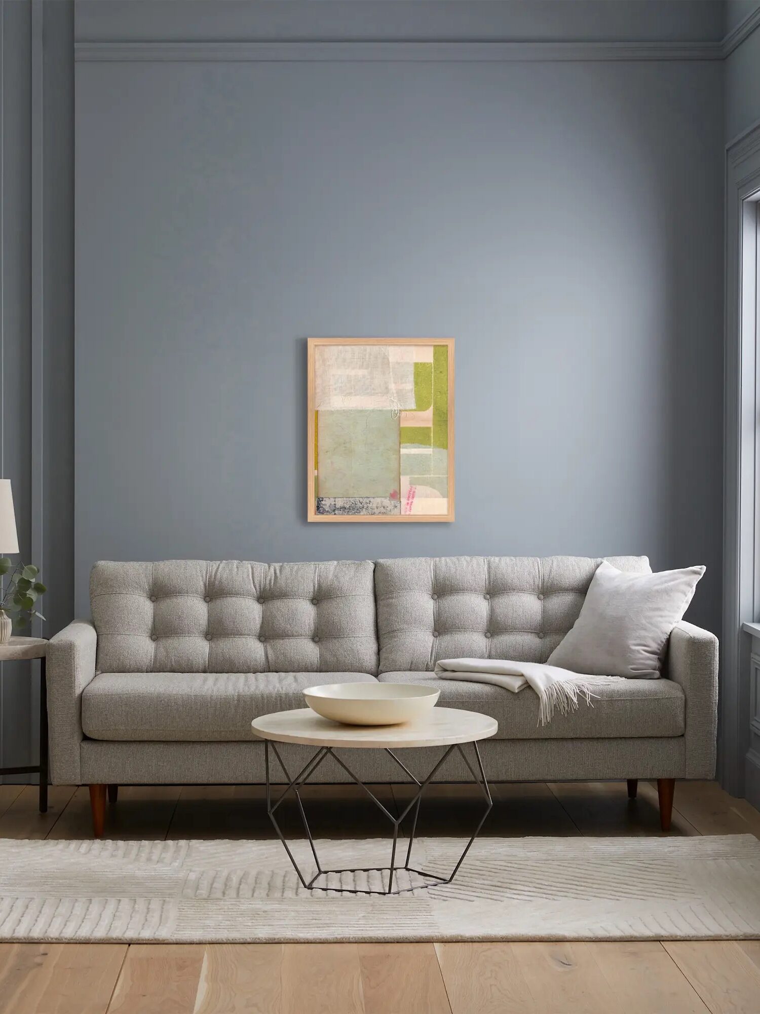 A modern living room with a light gray tufted sofa, white pillow and throw, round coffee table, abstract wall art, and a lamp on a side table against a pale blue wall.