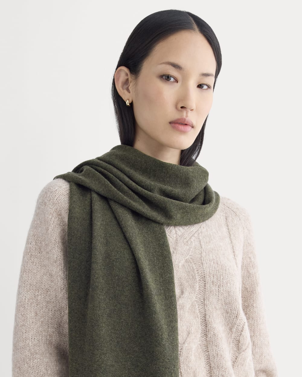 Woman with straight black hair wearing a light beige cable-knit sweater and an olive green scarf, standing against a plain light background.