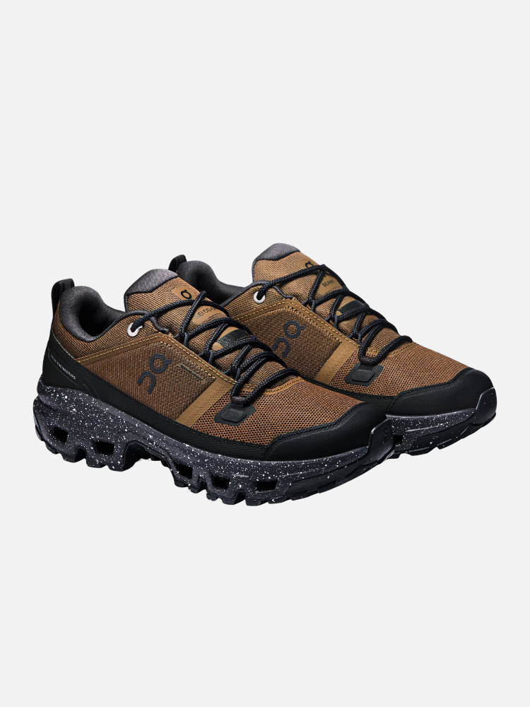 A pair of brown trail running shoes inspired by mens boots, with black soles and laces, featuring a textured upper and speckled midsoles on a white background.