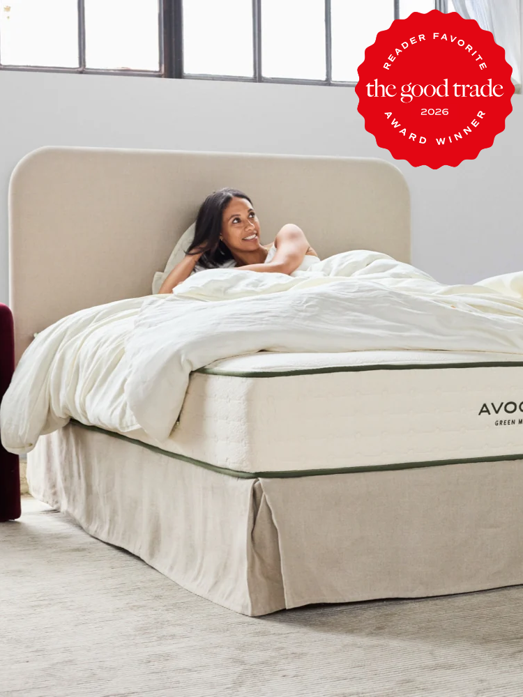 A woman smiles while sitting up in bed on a mattress labeled “Avocado,” with white bedding in a bright, modern bedroom. A red award badge is visible in the top right corner.