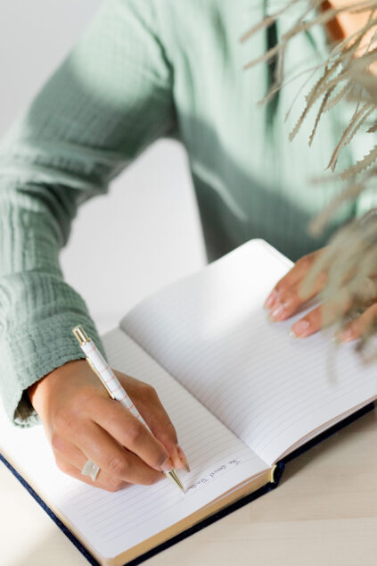 A person in a green long-sleeve shirt writes in a lined notebook with a pen, sitting at a light-colored table.