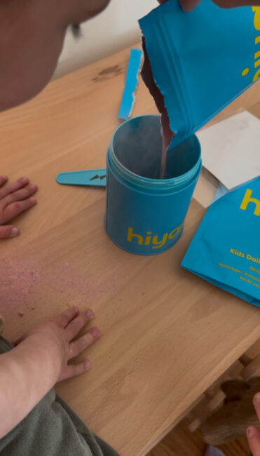 A person pours pink powder from a blue Hiya packet into a matching canister on a wooden table, with some powder spilled and a child's hands nearby.